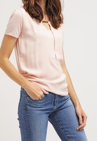 Frau in leichter pinkfarbener Kurzarmbluse mit Ausschnitt und blauen Jeans, eine Hand in der Tasche, vor einem schlichten hellen Hintergrund.