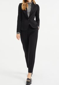 Černý na míru šitý oblek s výraznými rameny, blejzr na jedno zapínání a odpovídající zúžené kalhoty. Pod tím černobíle pruhovaný top.