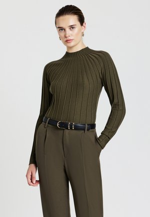 Donna che indossa un top a maniche lunghe ribbed di colore verde oliva, infilato in pantaloni abbinati con una cintura nera, in piedi su uno sfondo chiaro e uniforme.