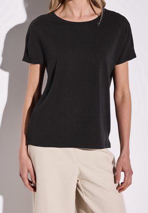 Frau trägt ein schlichtes schwarzes T-Shirt mit kurzen Ärmeln und hellbeige Hose vor neutralem Hintergrund.