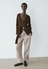 Vrouw draagt een bruin geribd vest met ceintuur, beige wijde broek, zwarte platte schoenen en een lange gouden hangersketting tegen een effen achtergrond.