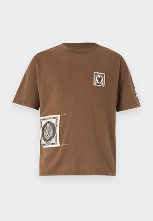 Rjava bombažna majica s krožnim izrezom, ki vsebuje dve okrasni nalepki, ki spominjata na vintage znamke v črni in beli barvi.