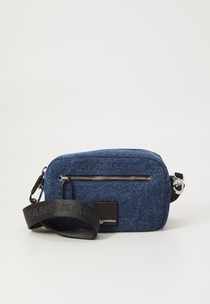 Blå denim crossbody-veske med glidelås, svart justerbar stropp og et rektangulært logo-patch. Strukturert stoff og sølvhardware.