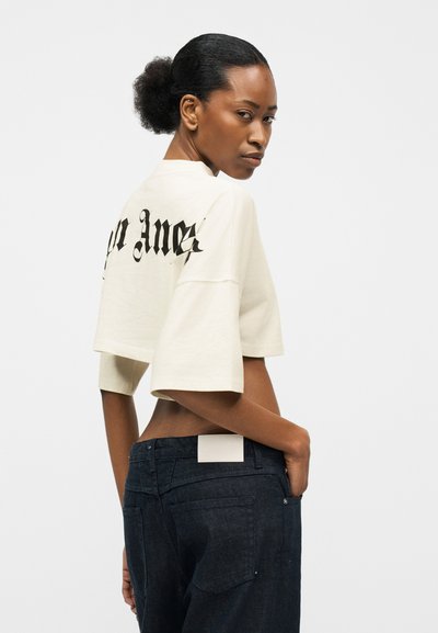 Γυναίκα με μαλλιά μαζεμένα προς τα πίσω που φοράει κομμένο λευκό t-shirt με μαύρο κείμενο και σκούρο τζιν ψηλόμεσο, κοιτάζοντας πάνω από τον ώμο της.