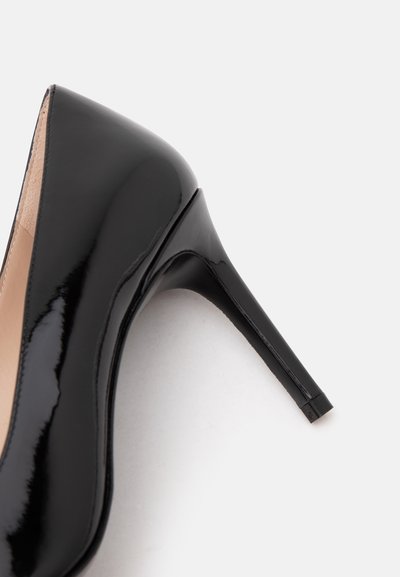Chaussure à talon haut en cuir verni noir avec un bout pointu et un talon aiguille fin sur fond blanc.