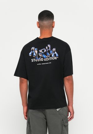 Svart t-skjorte med korte ermer som har en blank, metallisk stil "ALPHA" grafikk i blått på baksiden, med "STUDIO EDITION" under.