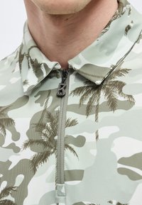 Homme portant une chemise camouflage vert clair avec des imprimés de palmiers marron et une courte fermeture éclair avec un tirette en forme de lettre "B".