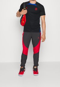 Czarna koszulka sportowa z niebieskimi akcentami, czerwoną logotypą, szare i czerwone spodnie z zakrzywionym wzorem oraz czerwone trampki z czarnymi detalami.