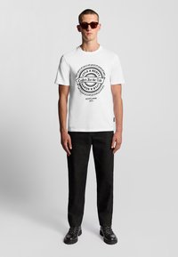 Camiseta blanca de algodón con un estampado gráfico circular negro que presenta el texto "Lyle & Scott" y "United for the Bold" en el frente. Pantalones negros y gafas de sol.