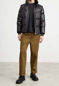 Svart läderpuffjacka med dragkedja och ribbade manschetter, kombinerad med bruna byxor och svarta kraftiga stövlar. Slät yta, modern design.