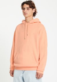 Hoodie pêche clair avec une poche avant, un capuchon à cordon de serrage et des poignets côtelés. Tissu doux avec un logo brodé minimaliste sur la poitrine.
