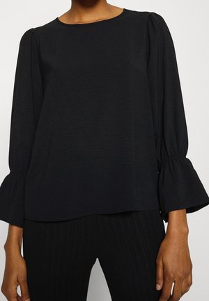 Blouse noire à manches longues avec poignets cloche froncés, col rond, portée avec un pantalon noir texturé, visible des épaules aux hanches.