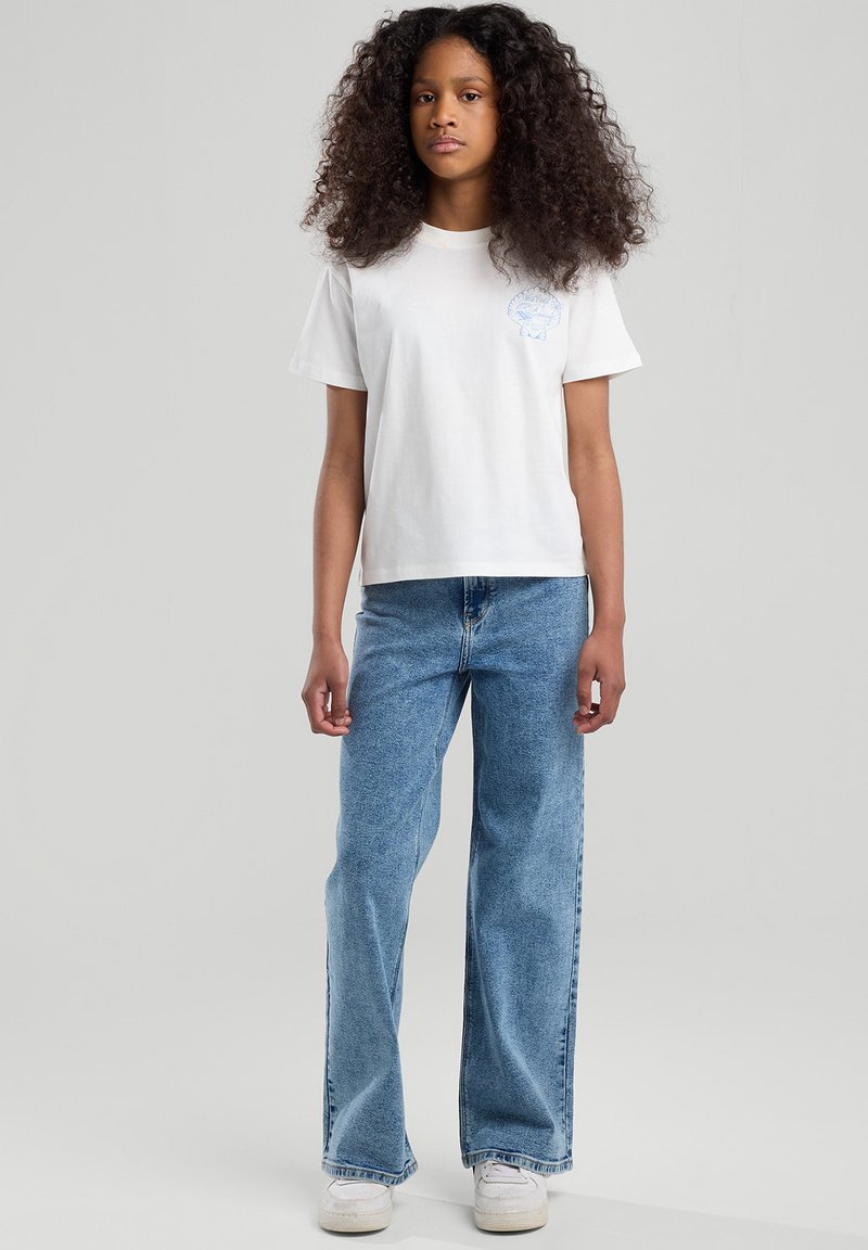 Valkoinen puuvillainen t-paita lyhyillä hihoilla ja vasemmassa rinnassa sininen graafinen kuvio, yhdistettynä vaaleansinisiin leveiden lahkeiden farkkuihin ja valkoisiin lenkkareihin.