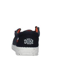 Scarpa in tessuto nero con parte superiore testurizzata, dettagli arancioni e suola bianca con macchie. Presenta una patch con logo che riporta la scritta "HEY DUDE" sul retro.