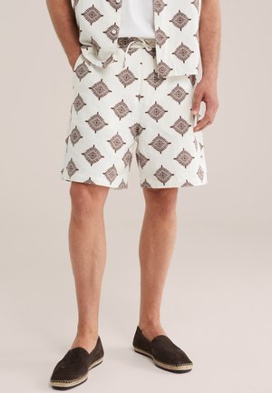 Mand iført hvide shorts og matchende skjorte med brunt geometrisk mønster, kombineret med mørkebrune slip-on sko.