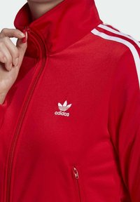Czerwona kurtka na zamek z wysokim kołnierzem, z białymi trzema paskami na rękawach oraz białym logo Adidas na piersi.