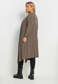 Brun randig lång cardigan med långa ärmar, öppen framsida och asymmetrisk nederkant, kombinerad med svarta kroppsnära byxor och kraftiga ankelstövlar.