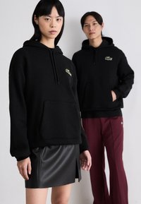Sudaderas con capucha negras y bolsillos canguro, con un logo verde. Un modelo lleva una falda de cuero negra, mientras que el otro lo combina con pantalones burdeos.