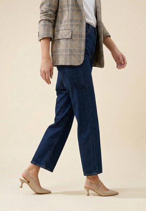 Persoon die loopt, gekleed in een beige geruite blazer, witte top, donkerblauwe spijkerbroek en beige slingback schoenen met kittenhak op een beige achtergrond.