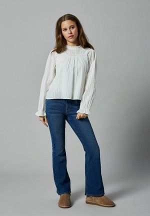 Blouse blanche à manches longues avec des plis verticaux et des poignets à volants, associée à un jean bleu évasé et des bottines beige. Fond neutre.