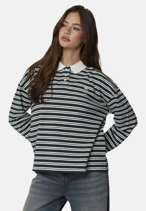 Giovane donna con capelli lunghi castani che indossa una polo a maniche lunghe a righe bianche e nere e jeans blu, posando con le mani sui fianchi.