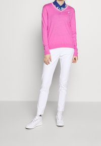 Růžový svetr s výstřihem do V, lehký materiál, dlouhé rukávy. Kombinováno s těsnými bílými kalhotami. Nošeno se bílými teniskami s nenápadným logem.