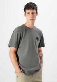 Grå bomulds t-shirt med rund halsudskæring, korte ærmer og et lille sort grafisk logo på venstre bryst. Blød tekstur, afslappet pasform.