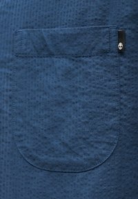 Gros plan sur une poche en tissu bleu texturé avec une petite étiquette noire portant un logo d'arbre blanc sur le bord supérieur droit.