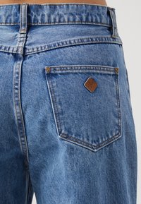 Jeans de mezclilla azul con cintura alta, un ligero desgaste y un parche de cuero marrón en el bolsillo trasero con forma de diamante.