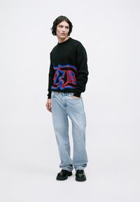 Maglione nero con design grafico blu e rosso, abbinato a jeans azzurri chiari e scarpe chunky nere. Il modello si trova su uno sfondo neutro.