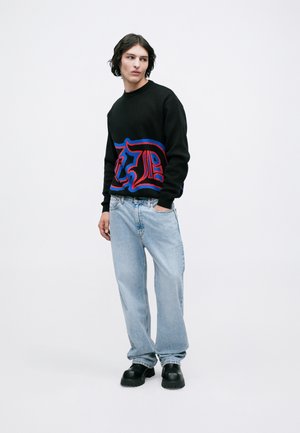 Sweater preto com design gráfico azul e vermelho, combinado com jeans azul claro e sapatos pretos de sola grossa. Modelo em pé contra um fundo neutro.