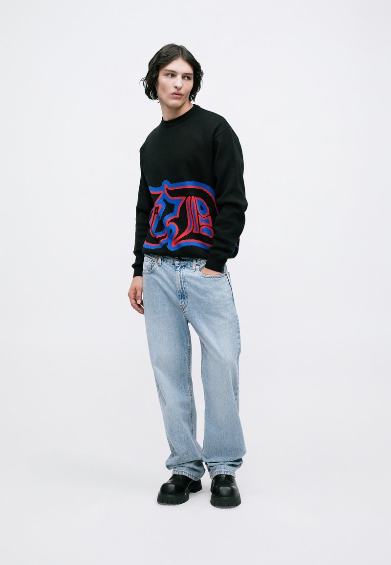 Maglione nero con design grafico blu e rosso, abbinato a jeans azzurri chiari e scarpe chunky nere. Il modello si trova su uno sfondo neutro.