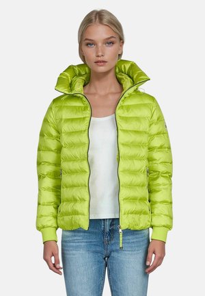 Frau trägt eine limettengrüne Steppjacke über weißem Hemd und blauer Jeans, steht vor einem schlichten grauen Hintergrund.