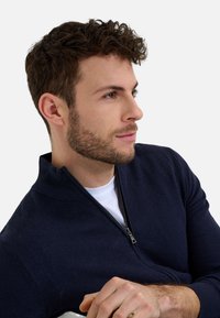Marineblauer Zip-Pullover mit strukturiertem Strick, Stehkragen und silbernem Reißverschluss. Getragen über einem weißen T-Shirt, mit einem Nahaufnahme-Profil.