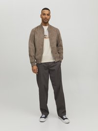 Giacca bomber in suede marrone sopra una t-shirt beige chiaro, abbinata a pantaloni grigio scuro e sneaker nere. Design semplice con polsini a coste.