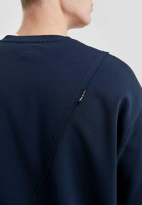Felpa blu navy con una particolare cucitura posteriore asimmetrica, caratterizzata da cuciture a vista e un piccolo etichetta tessuta vicino al colletto.