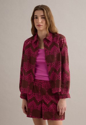 Frau trägt braune Jacke und Shorts mit magentafarbenem Blumenmuster und ein einfarbiges magentafarbenes Tanktop, steht vor einfarbigem Hintergrund.