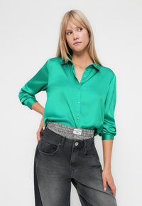 Camisa verde em cetim, com botões e mangas longas, combinada com jeans cinza escuro que têm uma cintura xadrez preta e branca.