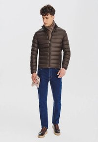 Brązowa kurtka puchowa z poziomymi szwami, zapinana na zamek z przodu i z wysokim kołnierzem. W połączeniu z niebieskimi dżinsami i brązowym obuwiem, trzyma beżowy szalik.