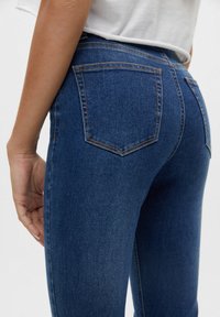 Tmavě modré denimové džíny s přiléhavým střihem, vybavené dvěma zadními kapsami a kontrastním prošitím podél švů. Hladká textura.