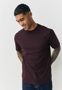 Bordo īsām piedurknēm t-krekls ar apaļu kakla izgriezumu, gluda tekstūra un pieguļošs dizains, valkāts kopā ar ziliem džinsiem.