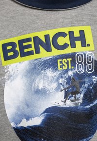 Camiseta gris con el audaz logo "BENCH." en amarillo y marino, con un gráfico azul de un surfista montando una ola, establecido en 1989.
