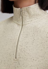 Zbliżenie na sweter z prążkami w odcieniu kremowym, w wielokolorowe plamki, z połowicznym zamkiem błyskawicznym w kolorze srebrnym, noszony przez osobę z krótkimi ciemnymi włosami.