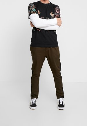 Man die met gekruiste armen staat, gekleed in een zwart bloemenpatroon korte mouwen shirt over een wit lange mouwen shirt, bruine broek en zwart-witte sneakers.