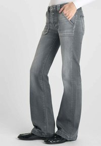 Jean gris évasé en denim avec poches avant, légère patine et fermeture éclair classique, associé à des chaussures noires brillantes.