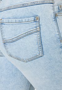 Světle modré džíny vyrobené z denimu, které mají zadní kapsu s kontrastním prošíváním a kovovým knoflíkem jako akcentem. Tkanina má mírně texturovaný vzhled.