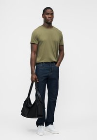 Bărbat purtând tricou verde măsline, blugi albastru închis, adidași albi, ținând o geantă neagră de umăr, stând pe un fundal alb simplu.