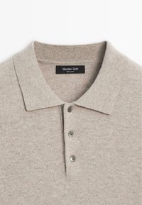 Polo beige realizzato in morbido tessuto a maglia, con colletto classico, patta con tre bottoni e una texture sottile. Etichetta del marchio visibile.