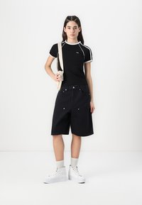 Svart kortärmad topp med vit kant, kombinerad med lösa svarta shorts. Modellen bär vita högklackade sneakers och har en beige axelväska.