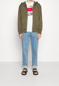 Sudadera con capucha de cremallera en verde oliva con bolsillos en la parte delantera, usada sobre una camiseta blanca con un gráfico rojo. Vaqueros azul claro remangados y sandalias negras.